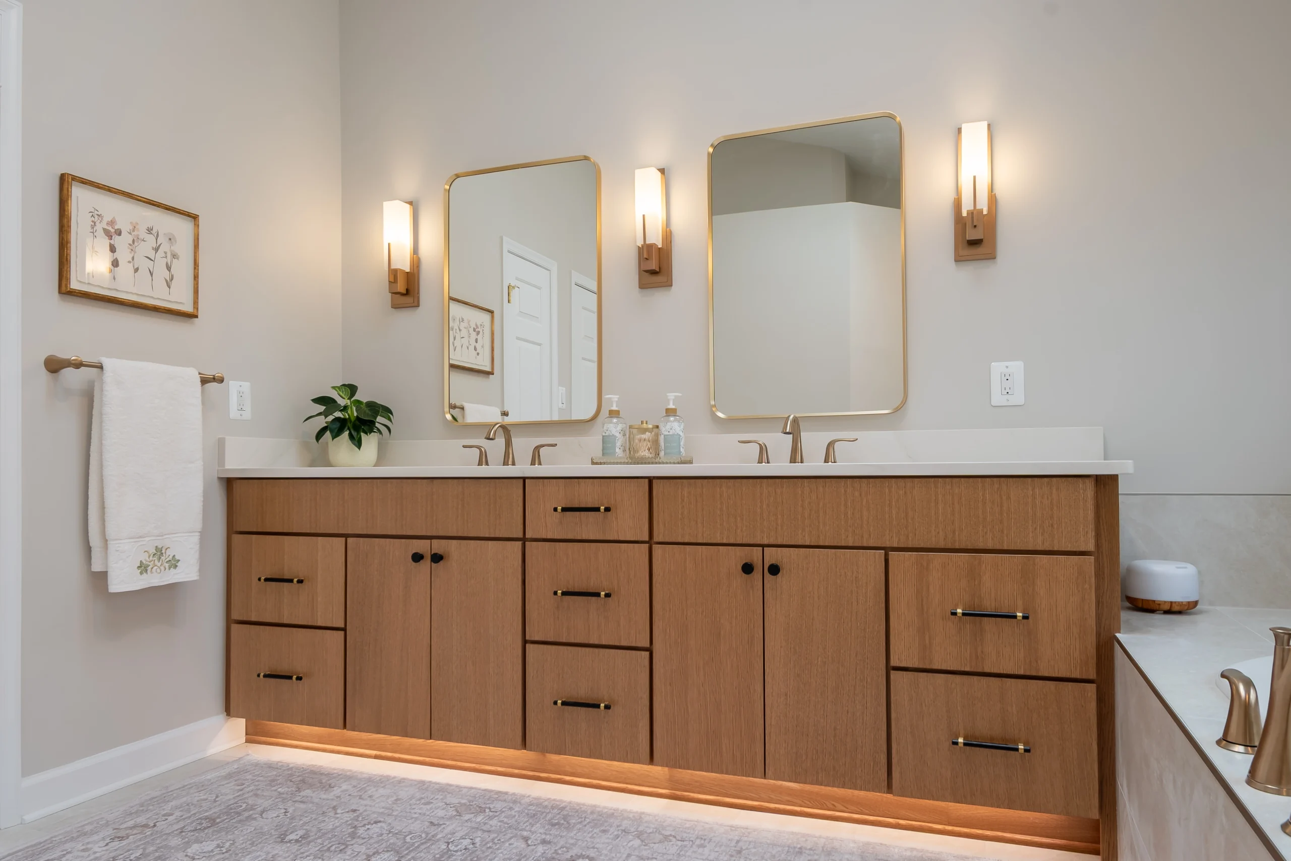Brown double vanity with a white counter, two gold-tone mirrors and lighting beneath the cabinets.