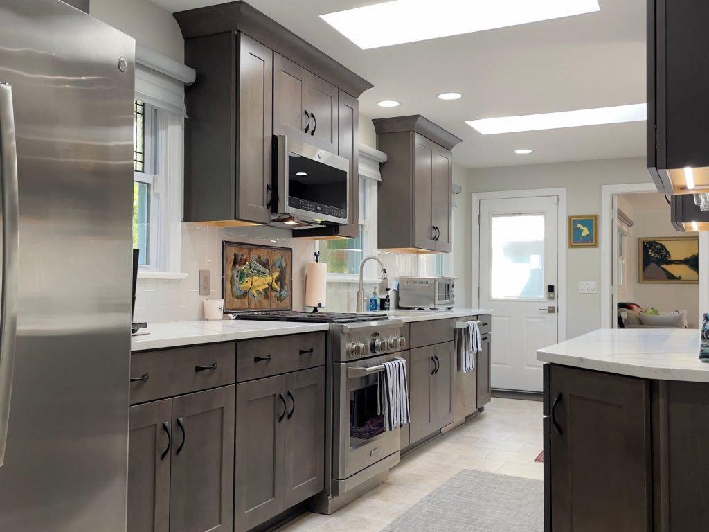 After: the same galley kitchen with dark gray shaker cabinets, white counters, a white tile backsplash and skylights overhead.