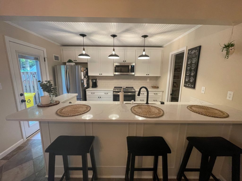 After: the same peninsula kitchen with white shaker cabinets, white veined counters, a subway tile backsplash, stainless appliances and the breakfast bar under the same three pendant lights.