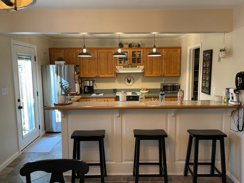 Before: peninsula kitchen with wood cabinets, gray counters, a white range and a tile-topped breakfast bar with three stools under three pendant lights.